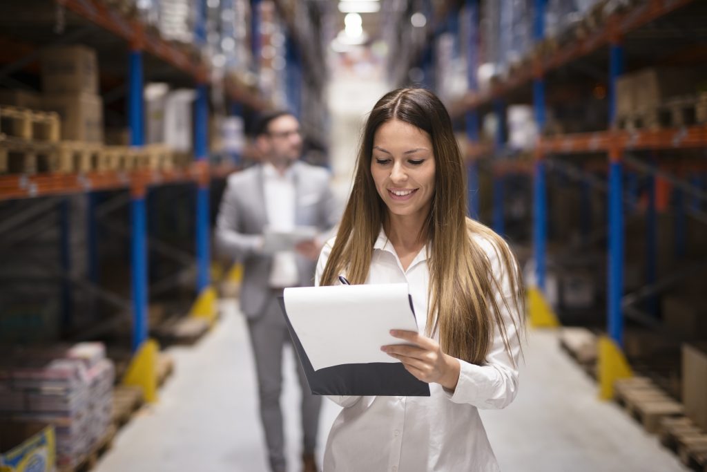 Portrait of gorgeous brunette woman manager controlling business in warehouse logistic center. Well dressed successful woman checking distribution while CEO walking behind her.